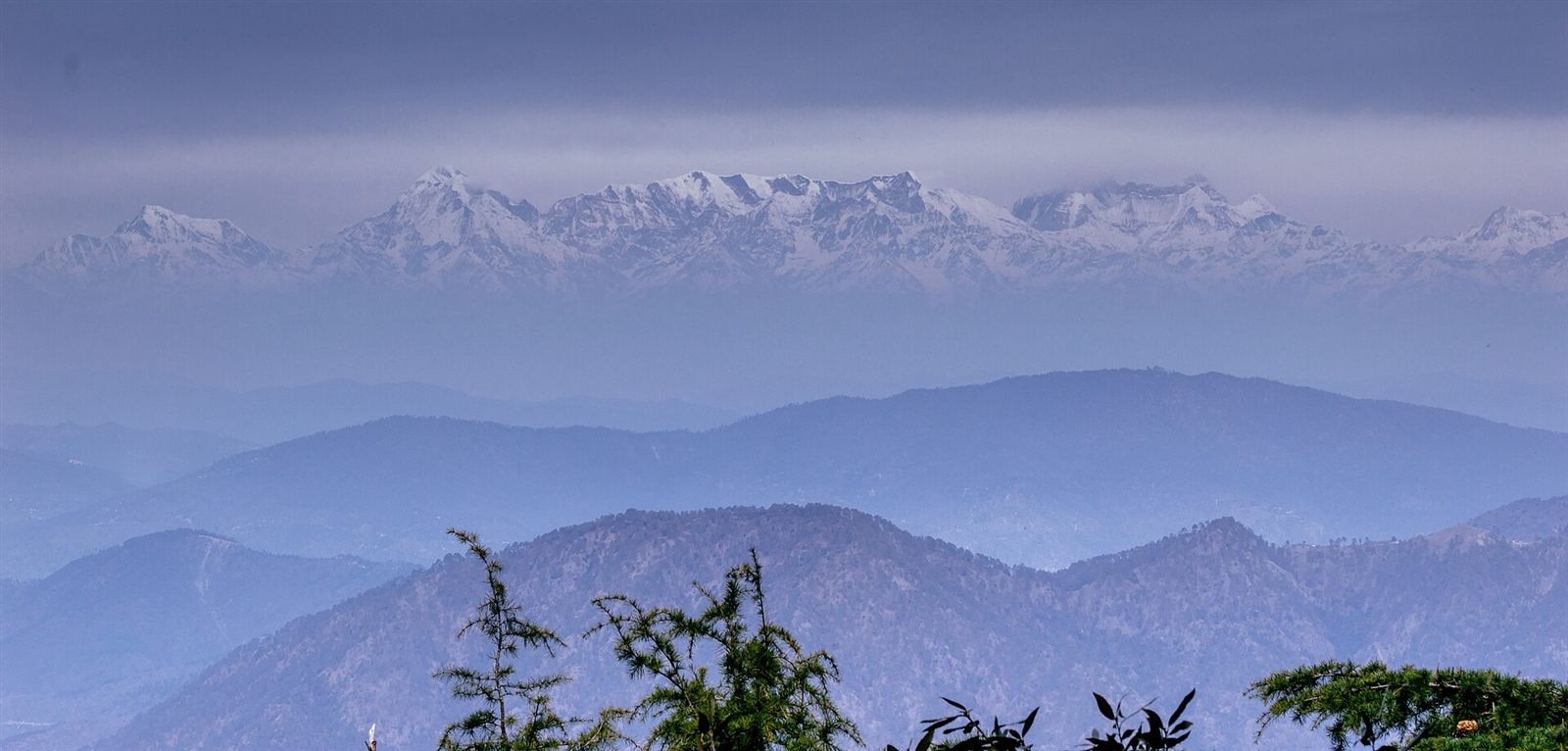 A long line of snow peaks above layers of blue ridges, seen from Himalaya Darshan point near Nainital